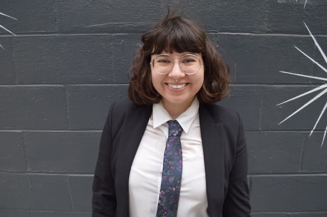A smiling young woman wearing a black suit and tie stands in front of a dark gray wall with white streaks.