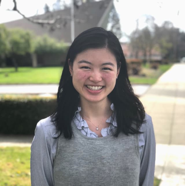 A young woman with long dark hair is smiling brightly in a park setting, with trees and buildings visible in the background.