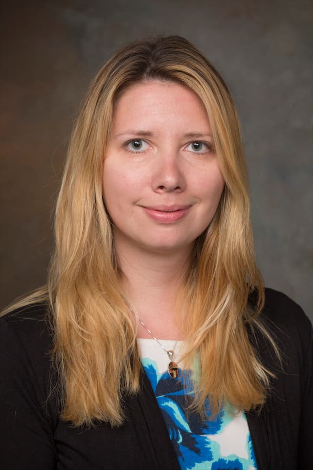 The image shows a woman with long, wavy blonde hair wearing a colorful floral blouse and a black jacket, smiling at the camera against a plain gray background.