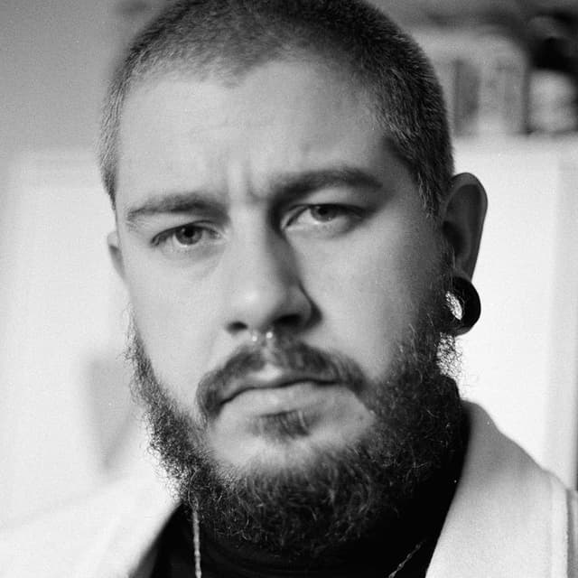 A close-up black and white portrait of a man with a beard, his gaze directed at the camera.