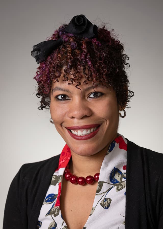 A smiling woman with curly, vibrant hair and a floral scarf against a plain background.
