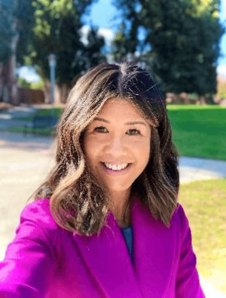 A smiling young woman with wavy brown hair stands in a park setting with trees and greenery in the background.