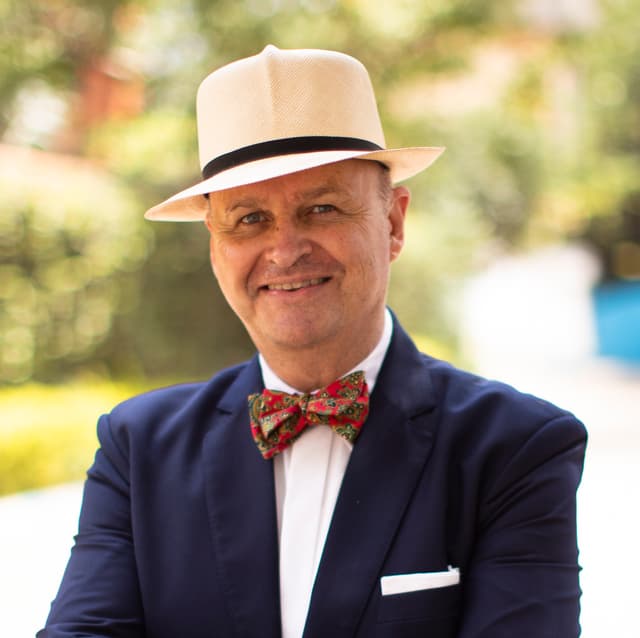 A well-dressed man wearing a navy blue suit, a red and white patterned bow tie, and a light-colored fedora hat stands in a lush, green outdoor setting.