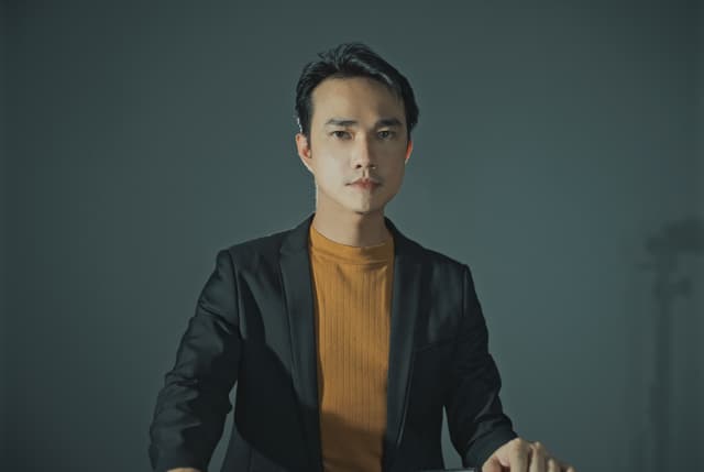 A man in a black jacket stands against a dark background, his expression serious and thoughtful.