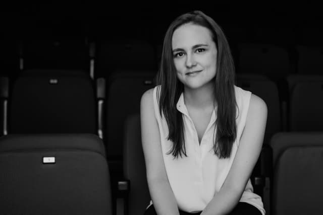 A young woman with long dark hair sits in a theater setting, her expression thoughtful and serene.