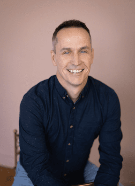 A smiling man with short dark hair wearing a navy blue button-down shirt against a light pink background.