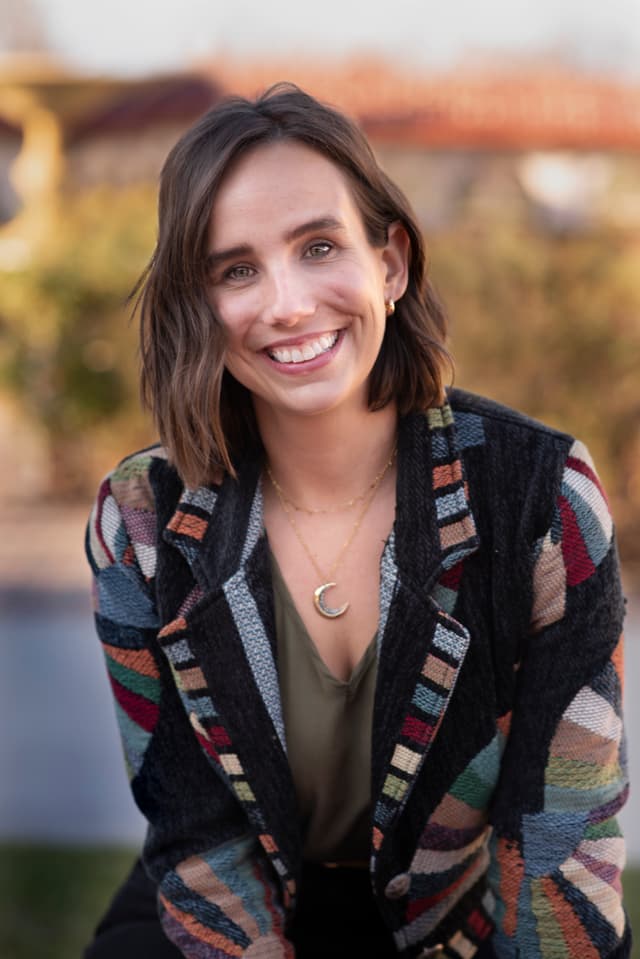 A smiling young woman with dark hair wearing a colorful patterned jacket stands in a scenic outdoor setting with a sunset sky in the background.