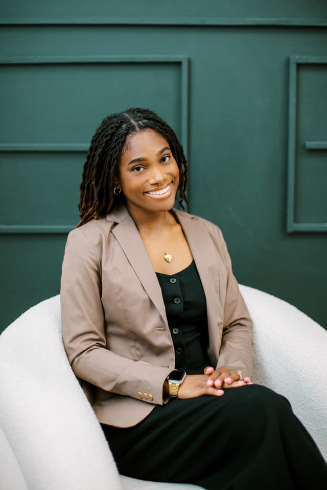 A smiling woman with dreadlocks sits comfortably in a white chair against a green wall background.