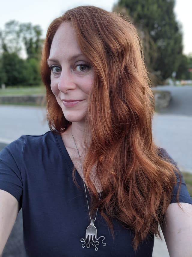 A young woman with long, wavy reddish-brown hair stands outdoors, smiling at the camera against a backdrop of trees and a body of water.
