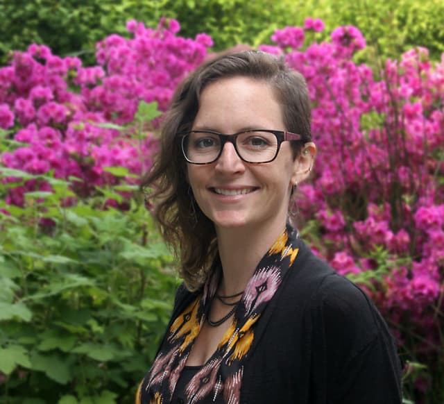 A smiling woman with glasses stands in front of a vibrant, blooming pink azalea bush in a lush, green outdoor setting.