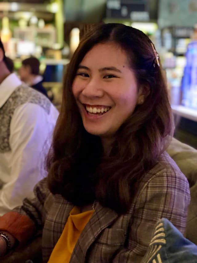A smiling young woman with long dark hair sits in a casual setting, surrounded by what appears to be a cafe or restaurant environment.
