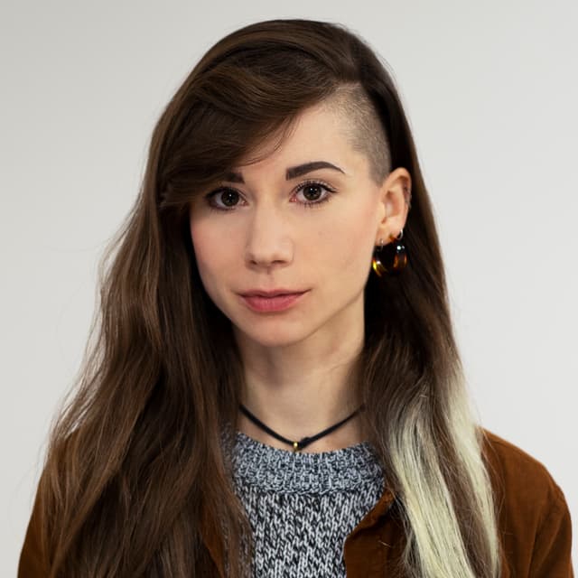 A young woman with long brown hair and a warm expression, wearing a gray sweater and brown jacket, stands against a plain white background.