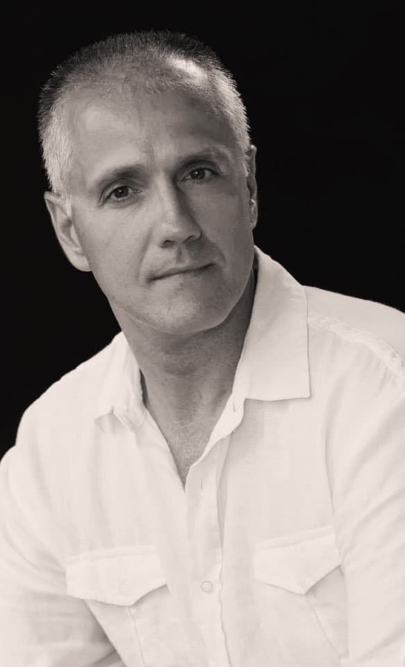 A black and white portrait of a middle-aged man with short gray hair and a serious expression, set against a dark background.