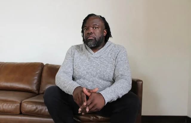A man with a beard wearing a gray sweater sits on a leather sofa in a room with a plain white wall.