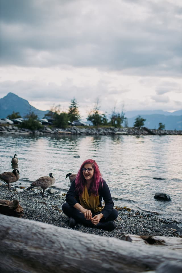 A person with vibrant purple hair sits cross-legged on a rocky shore, surrounded by a serene lake and mountains in the distance under an overcast sky.