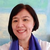 A smiling woman with short dark hair wearing a purple blouse stands in front of a blurred blue and green background.