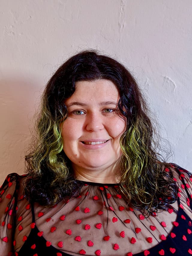 A smiling woman with curly dark and green hair, wearing a patterned blouse, stands in front of a plain white wall.