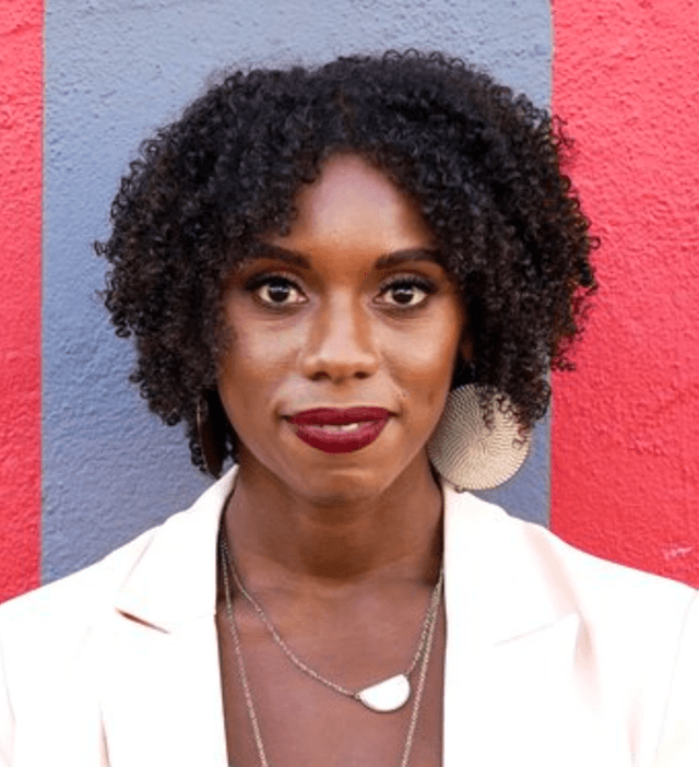 A woman with curly dark hair and a warm smile stands in front of a vibrant red wall, creating a striking visual contrast.