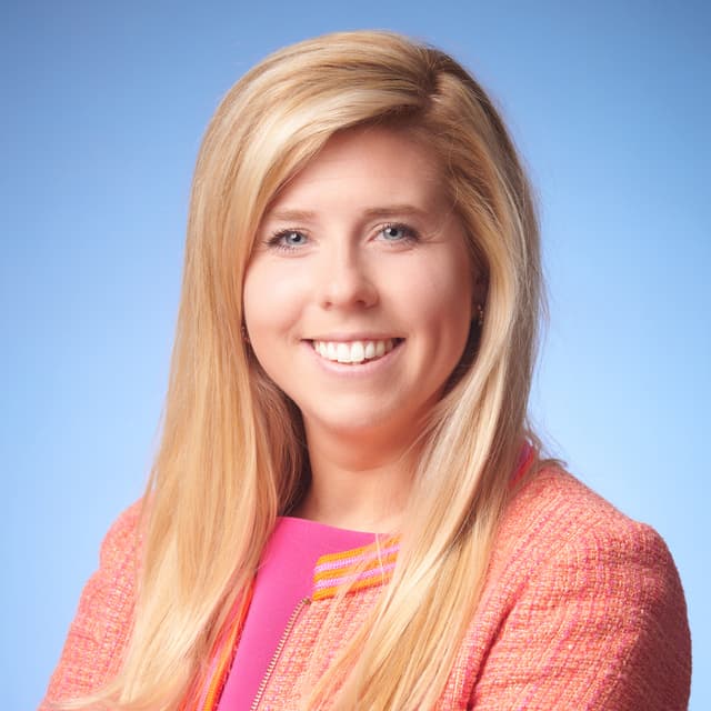 A smiling woman with long blonde hair wearing a pink top and a coral-colored jacket stands against a bright blue sky background.