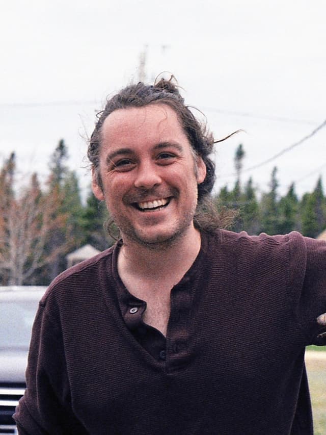 A smiling man with curly hair stands in front of a snowy forest landscape.