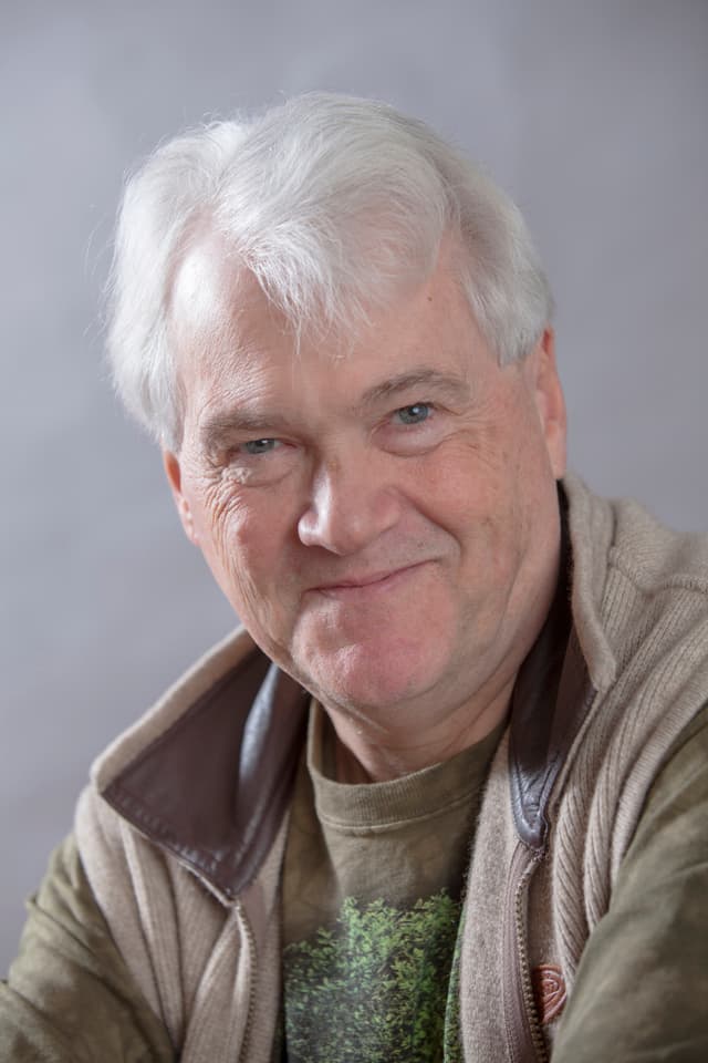 A smiling elderly man with white hair and a warm expression, wearing a beige jacket and sweater, set against a plain gray background.