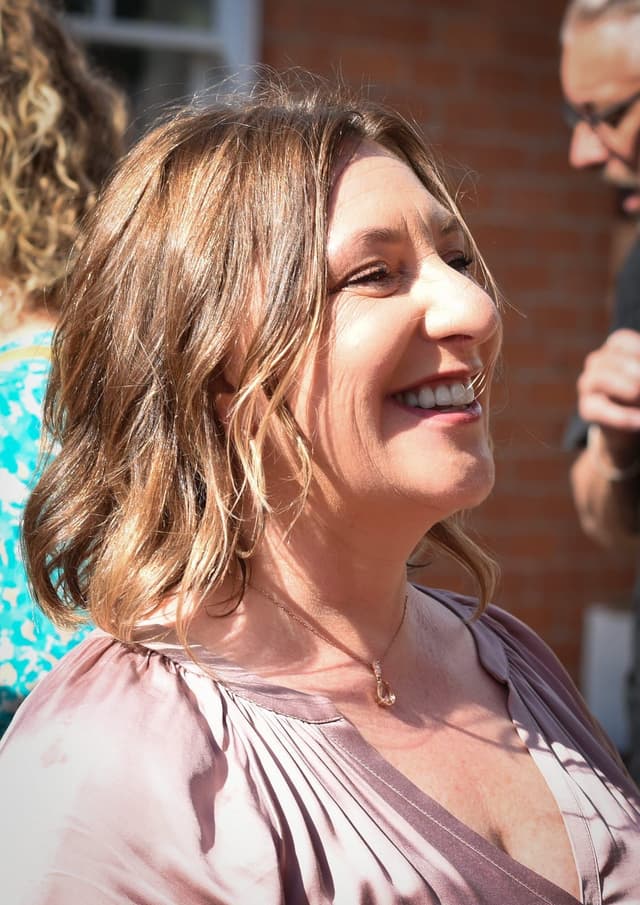 A smiling woman with wavy blonde hair stands in front of a brick wall, wearing a pink blouse and appearing to be in a cheerful mood.
