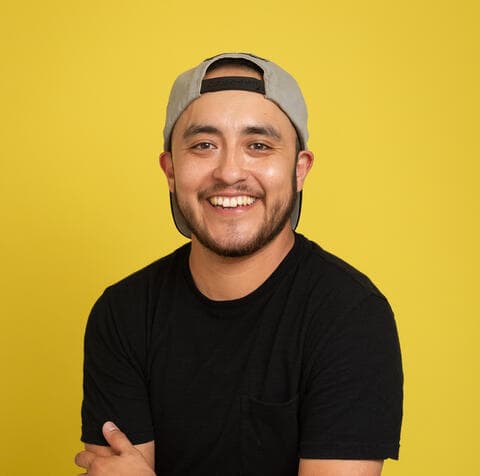 A smiling young man wearing a black t-shirt and a gray cap stands against a vibrant yellow background.