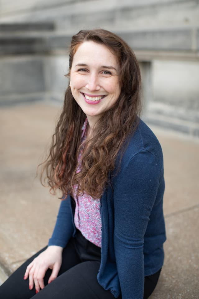 A young woman with curly brown hair is smiling brightly, seated on a concrete surface with a blurred background.