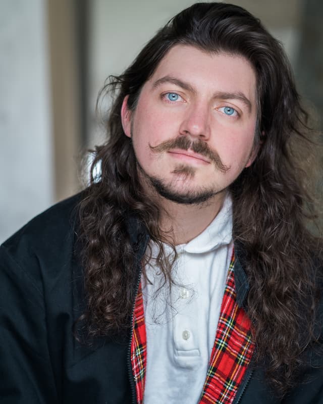 The image shows a man with long, curly dark hair and a mustache wearing a white shirt and a red and black plaid jacket, standing in what appears to be an indoor setting with a plain background.