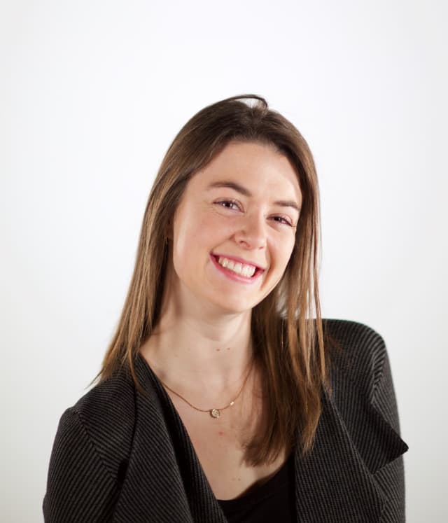 A smiling young woman with long brown hair wearing a black sweater stands against a plain white background.