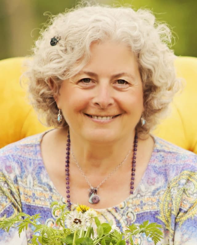 A smiling elderly woman with curly white hair wearing a colorful floral blouse sits in a lush, green outdoor setting.